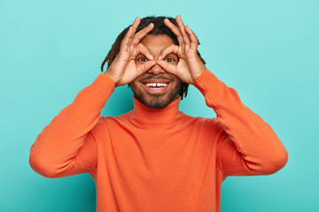 Wall Mural - Funny cheerful guy holds hands near eyes, pretends looking through binoculars tries to find love of life, has dreadlocks wears orange turtleneck, shows zero or okay gesture over eyes, isolated on blue