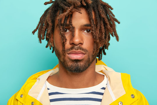 Close up portrait of serious dark skinned hipster with drealocks, has full lips, beard and mustache, wears striped jumper and yellow raincoat, listens attentively interlocutor, isolated on blue wall