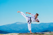 Karate man in a kimono performs a side leg-foot kick(Mae-geri) while standing on the green grass on top of a mountain.