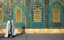 The Blue Mosque In Mazar-i-Sharif, Balkh Province In Afghanistan. Two Women Wearing White Burqas (burkas) Walk Past A Wall Of The Mosque Adorned With Colorful Tiles And Mosaics. Northern Afghanistan.