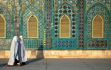 The Blue Mosque in Mazar-i-Sharif, Balkh Province in Afghanistan. Two women wearing white burqas (burkas) walk past a wall of the mosque adorned with colorful tiles and mosaics. Northern Afghanistan.