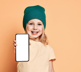 Smiling little girl kid in green modern winter hat showing white blank screen for text space of new popular mobile phone