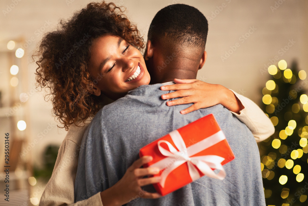 Thankful afro woman with christmas gift hugging husband at home - obrazy, fototapety, plakaty 