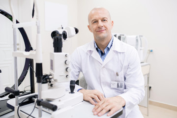 Poster - Happy bald mid-aged ophthalmologist in whitecoat sitting by workplace