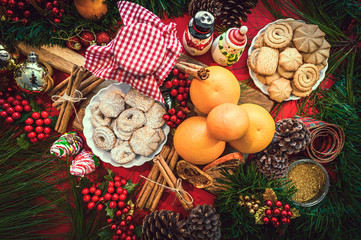 Christmas ingredients table with cookies, jam, oranges, tangerines, cinnamon, pine. Festive pine decoration on red rustic background, top view