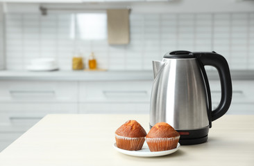 Poster - Modern electric kettle and muffins on wooden table in kitchen. Space for text