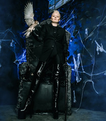 Photo of a female witch queen holding bird and sitting on a gothic scary black throne