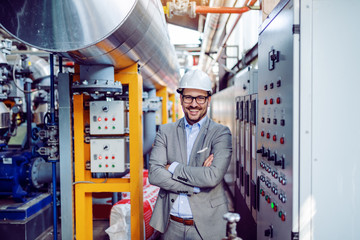 Smiling handsome caucasian businessman in gray suit and with helmet on head standing with arms folded next to dashboard. Power plant interior.