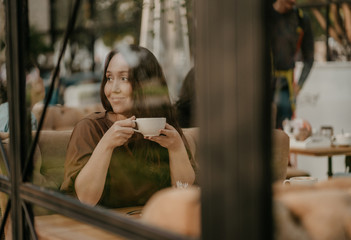 Charming brunette woman with long curly hair sitting at the window in cafe with cup of coffee in hands