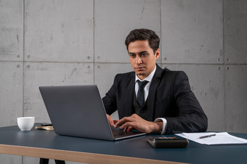 Portrait of serious and concentrated young man in dark suit typing on laptop in loft office. Concept of management and computer science