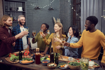 Wall Mural - Multi-ethnic group of smiling young people enjoying dinner together standing at table in modern interior and toasting with wine glasses, copy space