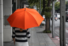 Back View Of Woman With Orange Umbrella Walking On A Street.