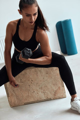 Wall Mural - Fitness young woman in black sportswear sit on fit box, dumbbells in hands.