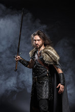 Man Dressed In Medieval Armor And Raincoat With Longs Word Over Smoke Background. Courage Fantasy Warrior Knight With Long Hair Concept Historical Photo