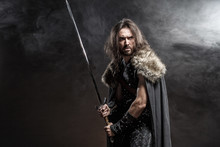 Man Dressed In Medieval Armor And Raincoat With Longs Word Fighting Against Enemy. Courage Fantasy Warrior Knight With Long Hair Concept Historical Photo