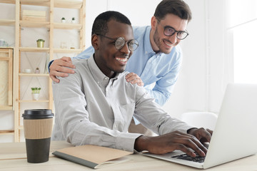 Friendly boss suppoting and explaining new task to smiling young employee, looking at laptop screen. Helping teacher consulting african american student