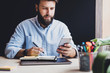 © foxyburrow - Bearded man doing work at desk. Male hipster holding smartphone and pen in his hands. Businessman taking notes, messaging on phone
