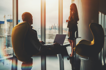 Two silhouettes of business partners on the top floor of a luxurious office skyscraper in front of the window with a construction site outside: man entrepreneur with a laptop and his female colleague