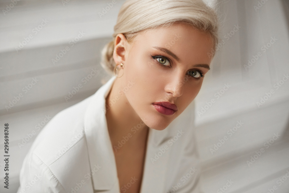 A fashionable young woman with perfect blond hair and perfect trendy makeup in an elegant white suit posing in studio. Beautiful blonde model girl with red full lips and deep seductive eyes. - obrazy, fototapety, plakaty 