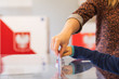 Woman's and kid's hands puting a card with a vote.to the ballot box during parliamentary elections. In the backround polish arms and flag