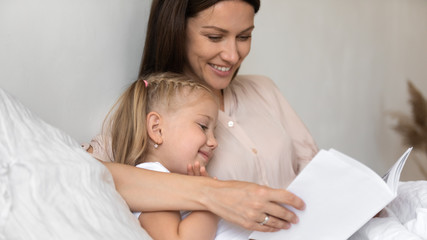 smiling mother reading book to cute little daughter in bed