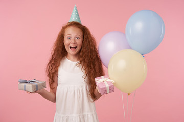 Wall Mural - Joyous female curly kid with long foxy hair being excited and surprised to get birthday present, happily looking in camera with gift box in hands over pink background. Children and celebration concept