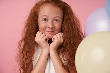 Wall Mural - Positive girl with red curly hair in white dress celebrates something, expresses true positive emotions, looking in camera cheerfully and leaning head on her hands, posing over pink background