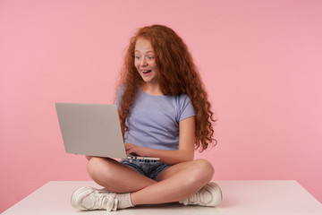 Wall Mural - Surprised cute female kid with curly long hair holding modern laptop and looking at screen cheerfully, keeping hands on keyboard while sitting with crossed legs over pink background