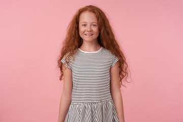 Wall Mural - Portrait of charming lond haired redhead girl with long hair wearing casual clothes, keeping hads along bod while standing over pink background, looking to camera with wide sincere smile