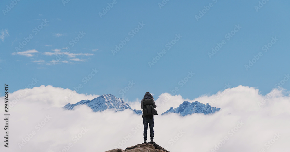 a man with backpack standing on mountain top, with snow mountains over clouds. Success, achievement and life goal concept - obrazy, fototapety, plakaty 