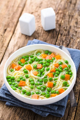 Wall Mural - Fresh homemade vegetable noodle soup with carrot, peas, onion and angel hair pasta in white soup bowl, spoon, salt and pepper on the side (Selective Focus, Focus one third into the image)