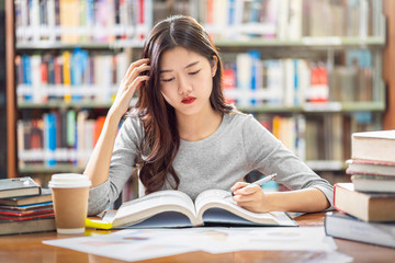 Wall Mural - Asian young Student in casual suit reading and doing homework in library of university or colleage with various book and stationary on the wooden table over the book shelf background, Back to school