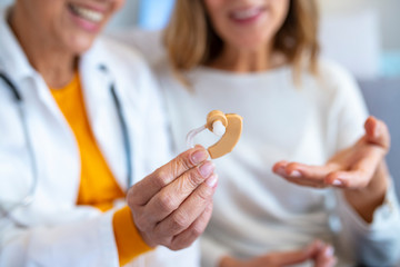 Mature female doctor hearing specialist in her office trying hearing aid equipment to a patient elderly senior woman. The doctor laryngologist explains to senior woman how to wear a hearing aid