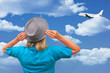 © Dmytro - Girl in hat looks at climbing aircraft that flies in clouds on blue sky. White middle aged woman stays back to us and touches her hat’s flaps. View with no face