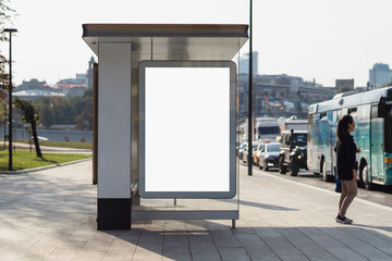 Bus stop with people standing by busy road with intensive traffic. Vertical billboard for commercial ads on side of station cabin. Public transport and private vehicles driving in lanes, traffic jam.