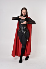 full length portrait of a pretty brunette woman wearing black leather fantasy costume with long red superhero cape. standing pose on a studio background.