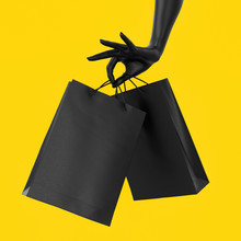 Close Up Black Female Hand Holding Black Paper Bag Isolated On Yellow Background. Shopping And Season Sale Concept Template Mock-up. Black Friday Concept. Copy Space. 3d Illustration.