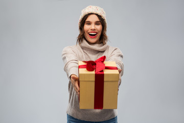 christmas, holidays and people concept - happy smiling young woman in knitted winter hat and sweater holding gift box over grey background