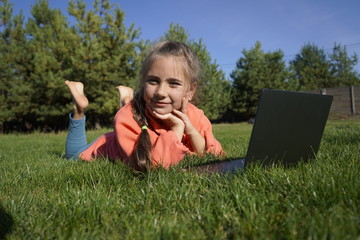 A girl of seven years old works on a laptop on the green grass. Distance learning. Girl with a laptop on a background of pines. Girl with a computer on the street.