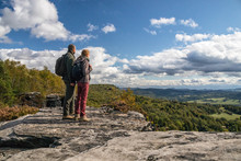Two Hikers (couple - Man And Woman) Up On The Rock - Looking Into Valley Below Mountain On Forest And Blue Sky With White Clouds