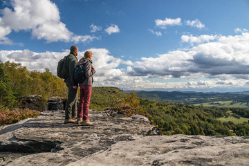 two hikers (couple - man and woman) up on the rock - looking into valley below mountain on forest and blue sky with white clouds