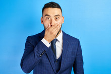 Frightened Shocked Asian Kazakh Man In Blue Suit And Tie With Bulging Surprised Eyes Closes Mouth With His Hand Isolated In Studio