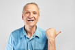 © amixstudio - Positive gray-haired senior citizen joyfully smiles, shows a finger to the side on free copy space isolated in white studio