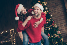 Photo Of Funky Couple Spending X-mas Eve In Decorated Garland Lights Room Carrying Piggyback Celebration Mood Wear Red Pullovers And Santa Hats Indoors