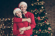 Photo of cute couple spending christmas time eve in decorated garland lights room standing piggyback near x-mas tree indoors wear red pullovers and santa hats