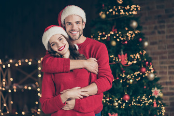 Wall Mural - Photo of cute couple spending christmas time eve in decorated garland lights room standing piggyback near x-mas tree indoors wear red pullovers and santa hats