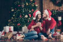 Photo Of Dreamy Couple Newyear Night In Decorated Garland Lights Room Sitting Cozy On Floor Near X-mas Tree Drinking Hot Beverage Indoors Wearing Red Sweaters And Santa Hats