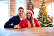 © Krakenimages.com - Young couple wearing santa claus hat sitting on chair and table around christmas tree at home showing and pointing up with fingers number four while smiling confident and happy.