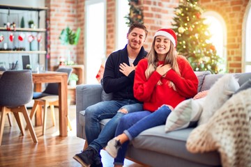 Wall Mural - Young couple wearing santa claus hat sitting on the sofa around christmas tree at home smiling with hands on chest with closed eyes and grateful gesture on face. Health concept.