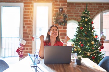 Beautiful woman sitting at the table working with laptop at home around christmas tree celebrating surprised and amazed for success with arms raised and eyes closed. Winner concept.